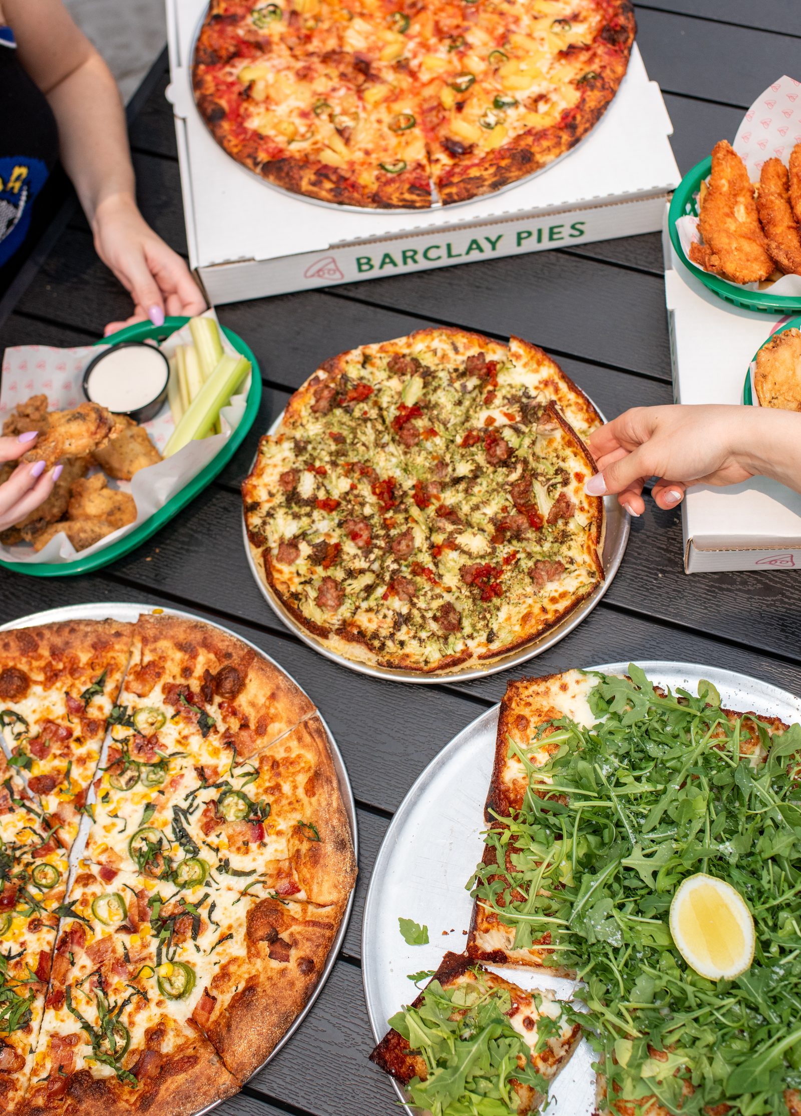 A spread of Barclay Pies pizza slices, wings, fries and salads in natural light